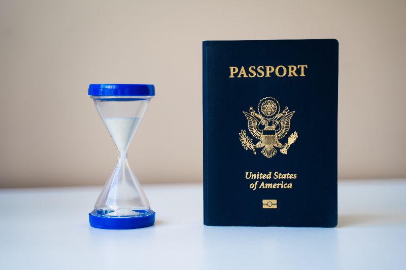 A blue U.S. passport standing next to an hourglass on a white table.