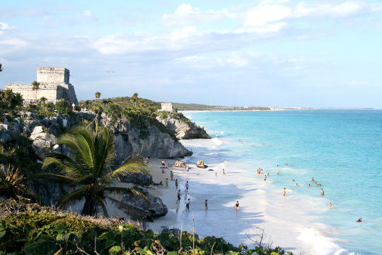 People playing on beach in Tulum Mexico with ruins at top of hill.